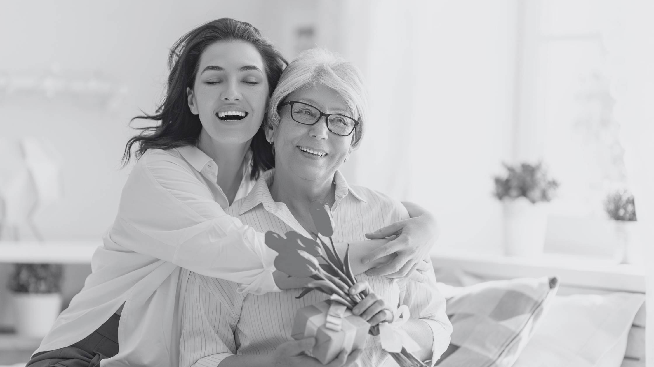 Black and white photo of a daughter hugging her mother who is holding a gift and flowers.