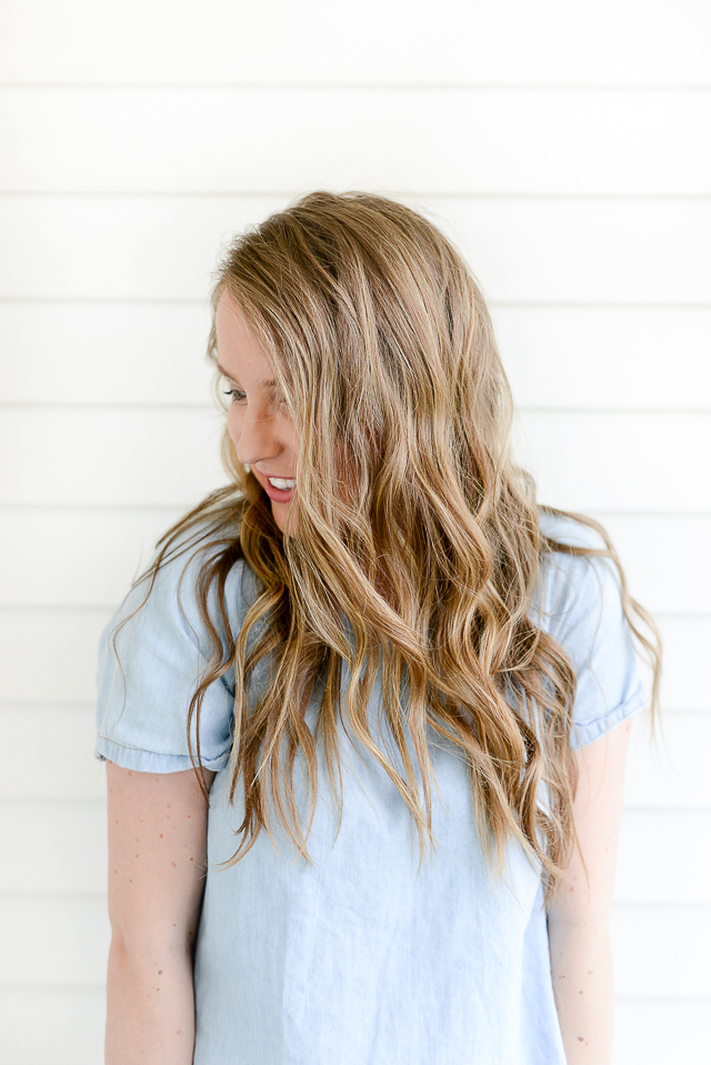 Woman with long, light-brown beachy waves looking to the side against a white shiplap wall.